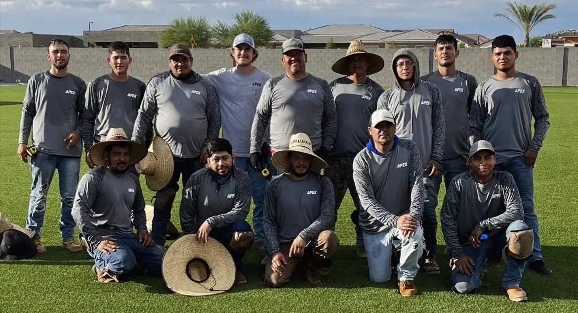 A group of sixteen men in matching long-sleeve shirts and hats pose together outdoors on grass, with a residential neighborhood and a concrete wall in the background.