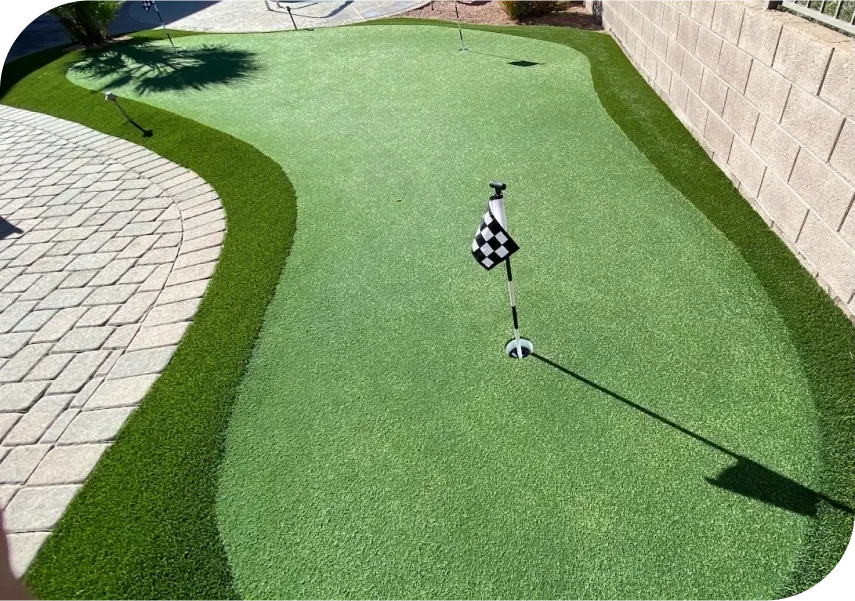 A backyard putting green with artificial turf, bordered by a lighter green strip and a stone patio. A hole with a checkered flag is in the center, and a brick wall runs along the right side. Shadows are cast on the green.