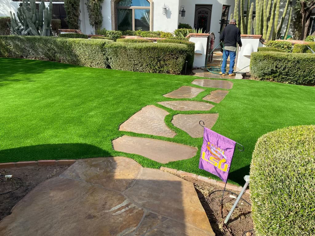 A landscaped yard with bright green artificial grass, a stone pathway by an expert Pavers Installer Texas, trimmed bushes, and an LSU flag. A person waters plants near the entrance of a white house in the background.