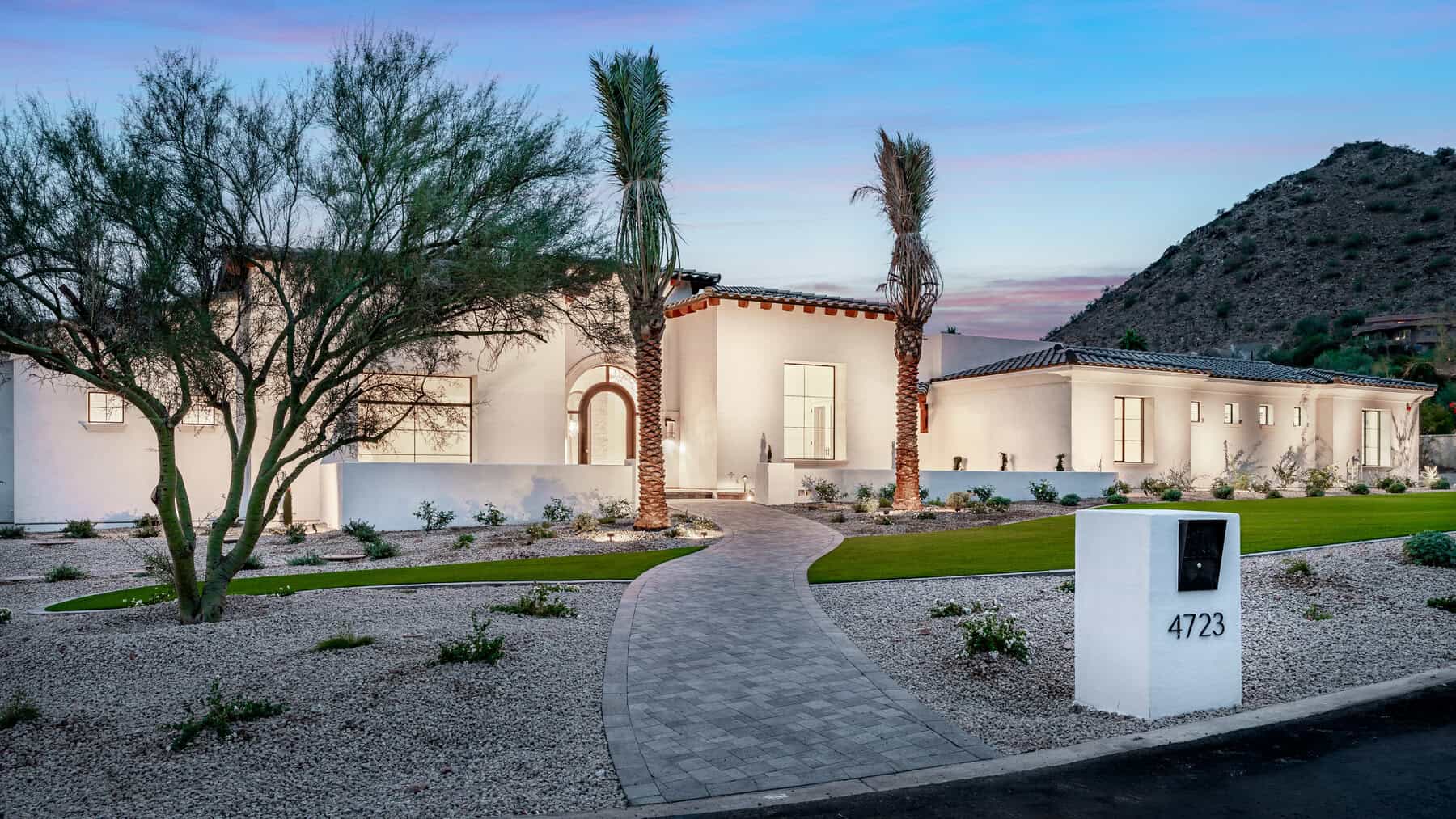 Modern white single-story house with large windows, palm trees, and desert landscaping at sunset. A curved stone pathway, expertly crafted by a Pavers Installer Texas, leads to the entrance; mailbox 4723 is visible in the foreground.