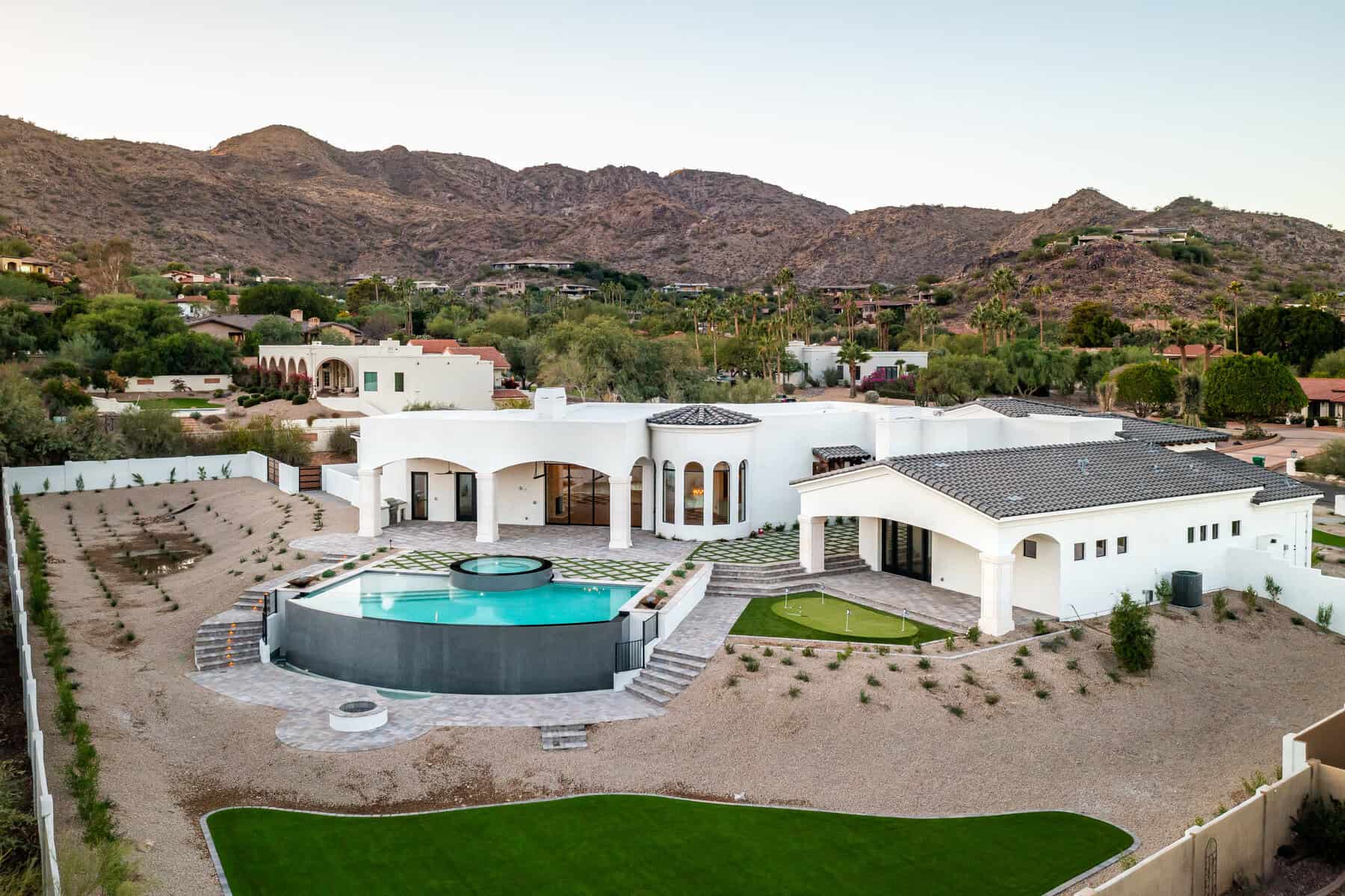 Aerial view of a large, modern white house with a circular swimming pool in the backyard, beautiful pavers installed by a top Pavers Installer Texas, surrounded by mountains, desert landscaping, and patches of green lawn.