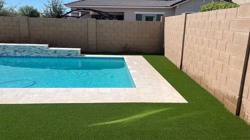 A backyard swimming pool with light-colored tile and expertly designed pavers installed by a trusted Pavers Installer Texas, artificial green grass along the edges, and tan brick walls enclosing the area. A house and trees are visible in the background.