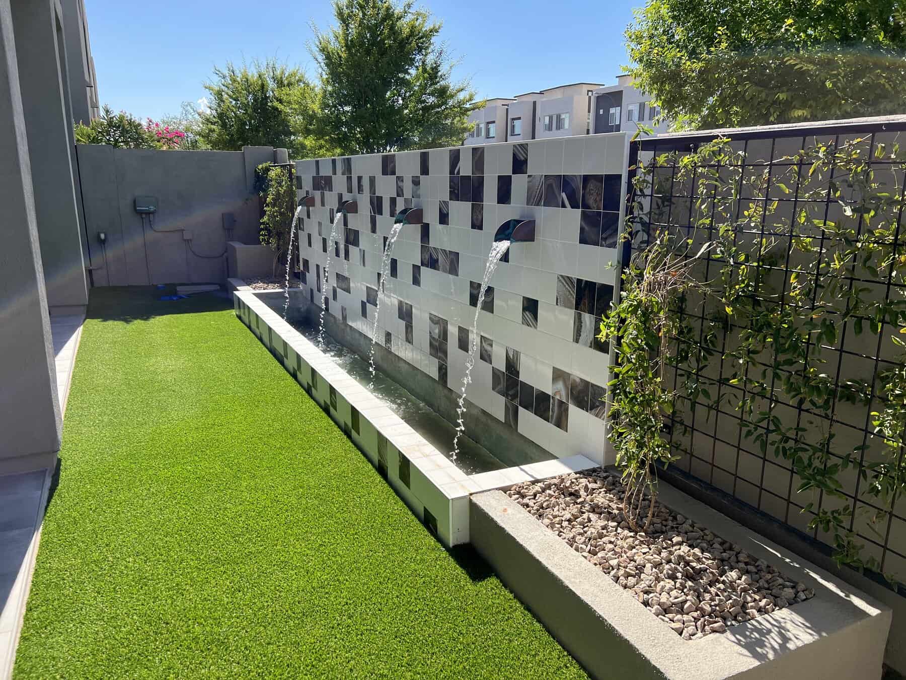 A modern backyard by a skilled Pavers Installer Texas features a tiled fountain wall with three water spouts, artificial turf, and trellises with climbing plants along the sides under a clear blue sky.