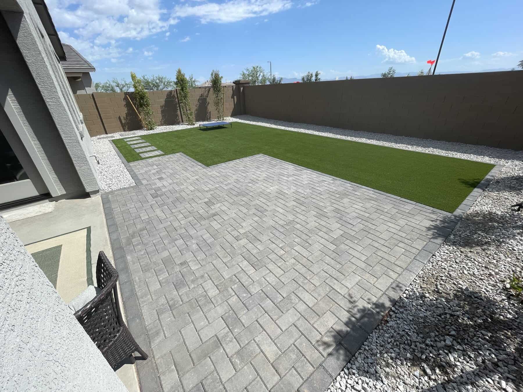 Modern backyard designed by a Pavers Installer Texas, featuring light gray pavers, a rectangular artificial grass lawn, white gravel borders, a low concrete wall, young trees along the fence, and a clear blue sky above.
