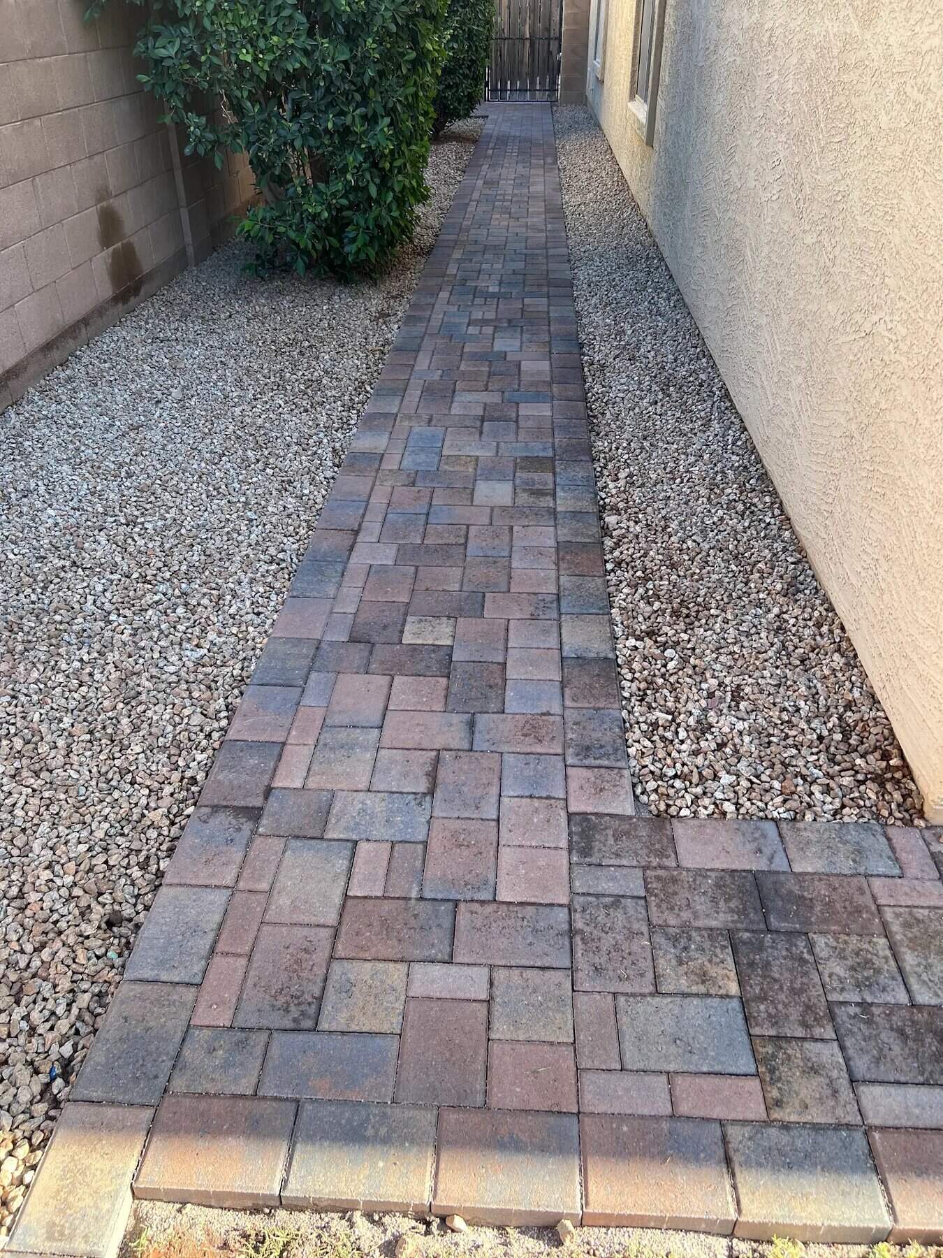 A narrow pathway made of rectangular pavers, expertly crafted by a Pavers Installer Texas, runs alongside a beige house, bordered by gravel on both sides and a bush near a block wall fence.
