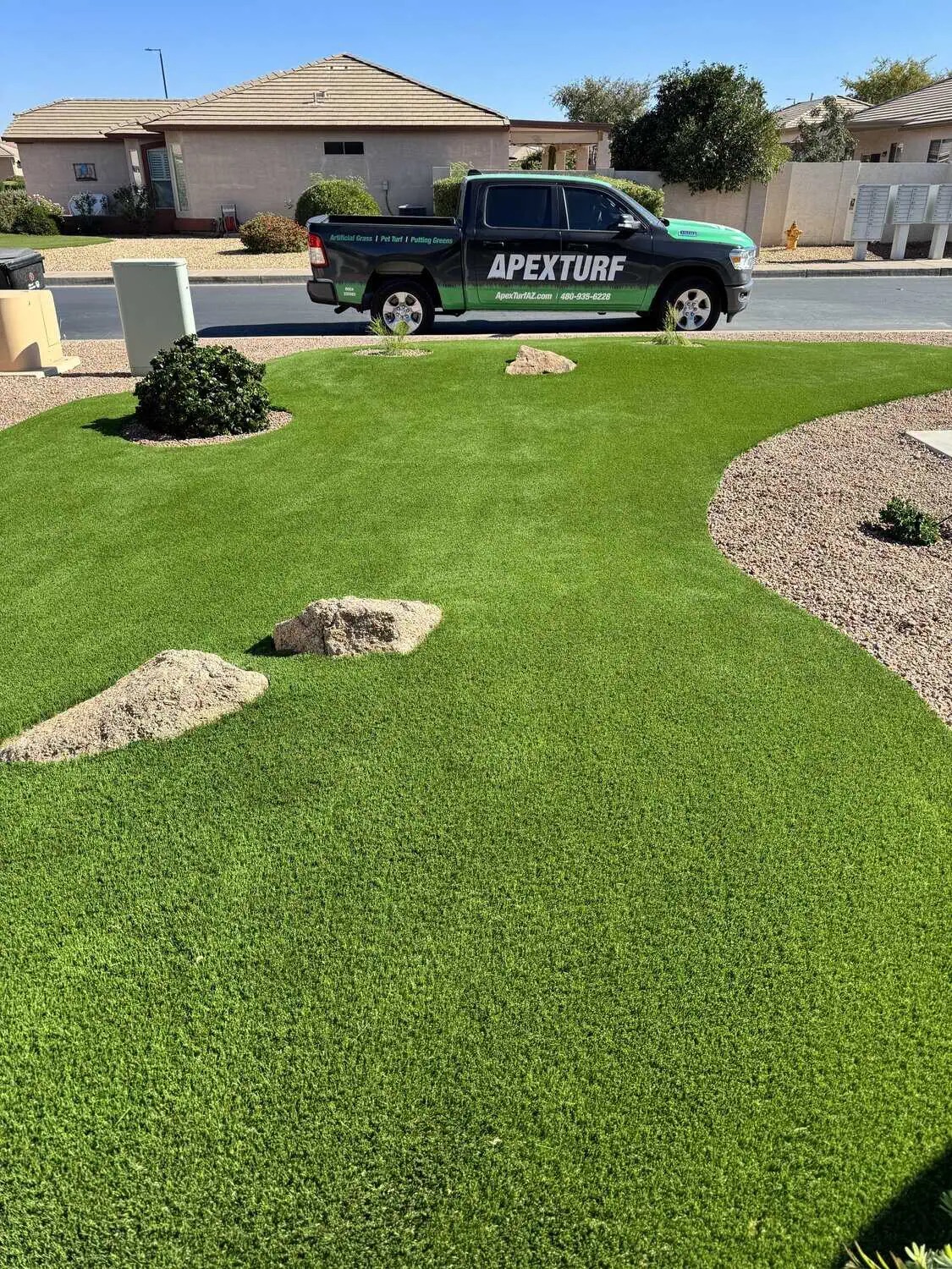 A neatly manicured artificial lawn, expertly installed by an Artificial Grass Installer, features small piles of dirt in a front yard. A green truck labeled "APEXTURF" is parked on the street in front of a house under a clear sky.