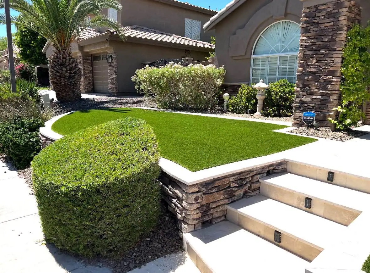 A neatly manicured front yard by an expert Artificial Grass Installer features artificial grass, stone retaining walls, trimmed bushes, a palm tree, and decorative landscaping in front of a beige house with arched windows and a tiled roof.