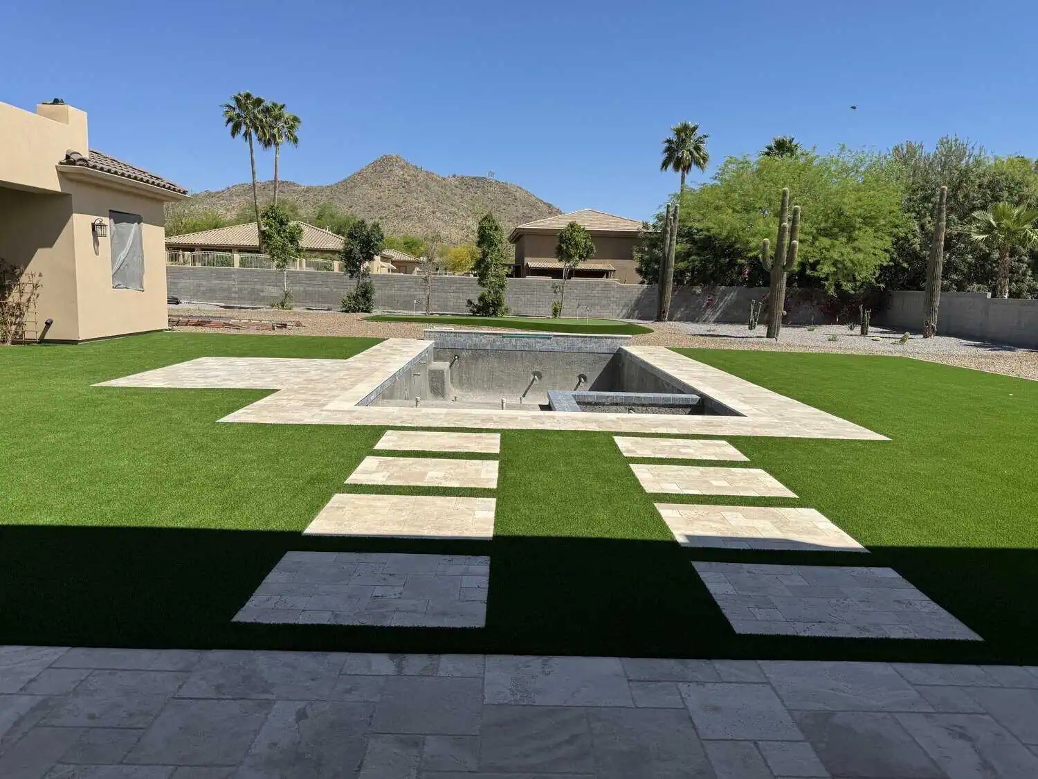 A backyard with green artificial turf installed by a professional Artificial Grass Installer, a rectangular unfinished pool surrounded by stone tiles, and a stone walkway. Houses, trees, and mountains stand in the background under a clear blue sky.