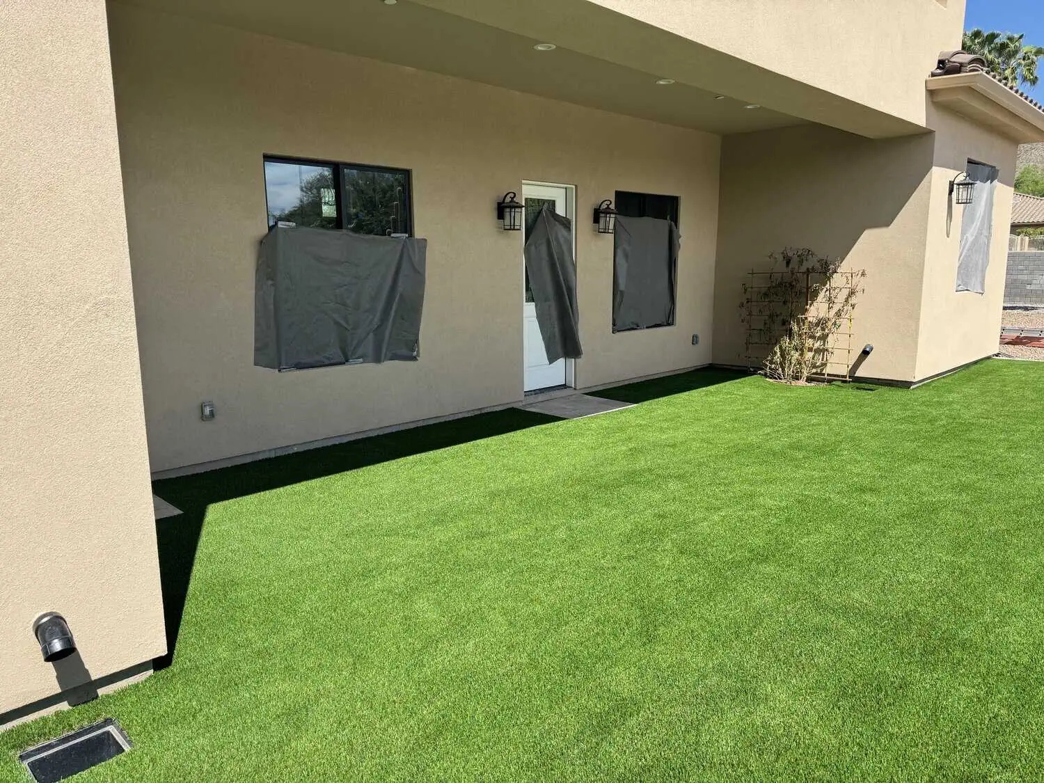 A backyard with bright green artificial turf installed by an expert artificial grass installer surrounds a beige house. Windows and a door are covered with gray material, and a small lattice with plants rests against the wall.