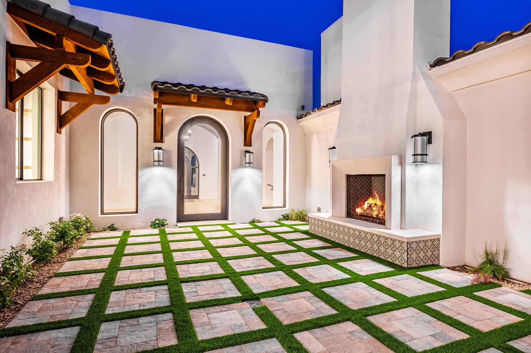 A backyard putting green with artificial turf, golf flags, large rocks, a white sand trap, and lush landscaping, all set against a tall tan wall and adjacent to a nearby patio area.