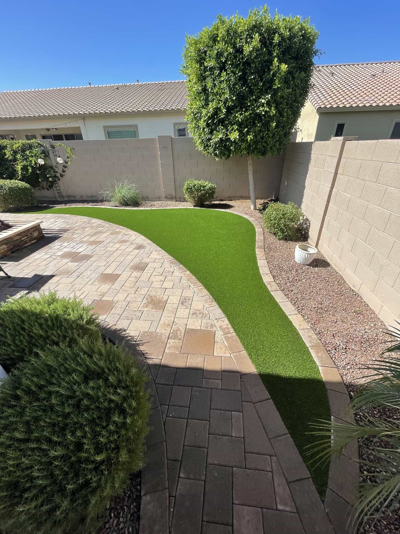 A backyard putting green with artificial turf, golf flags, large rocks, a white sand trap, and lush landscaping, all set against a tall tan wall and adjacent to a nearby patio area.