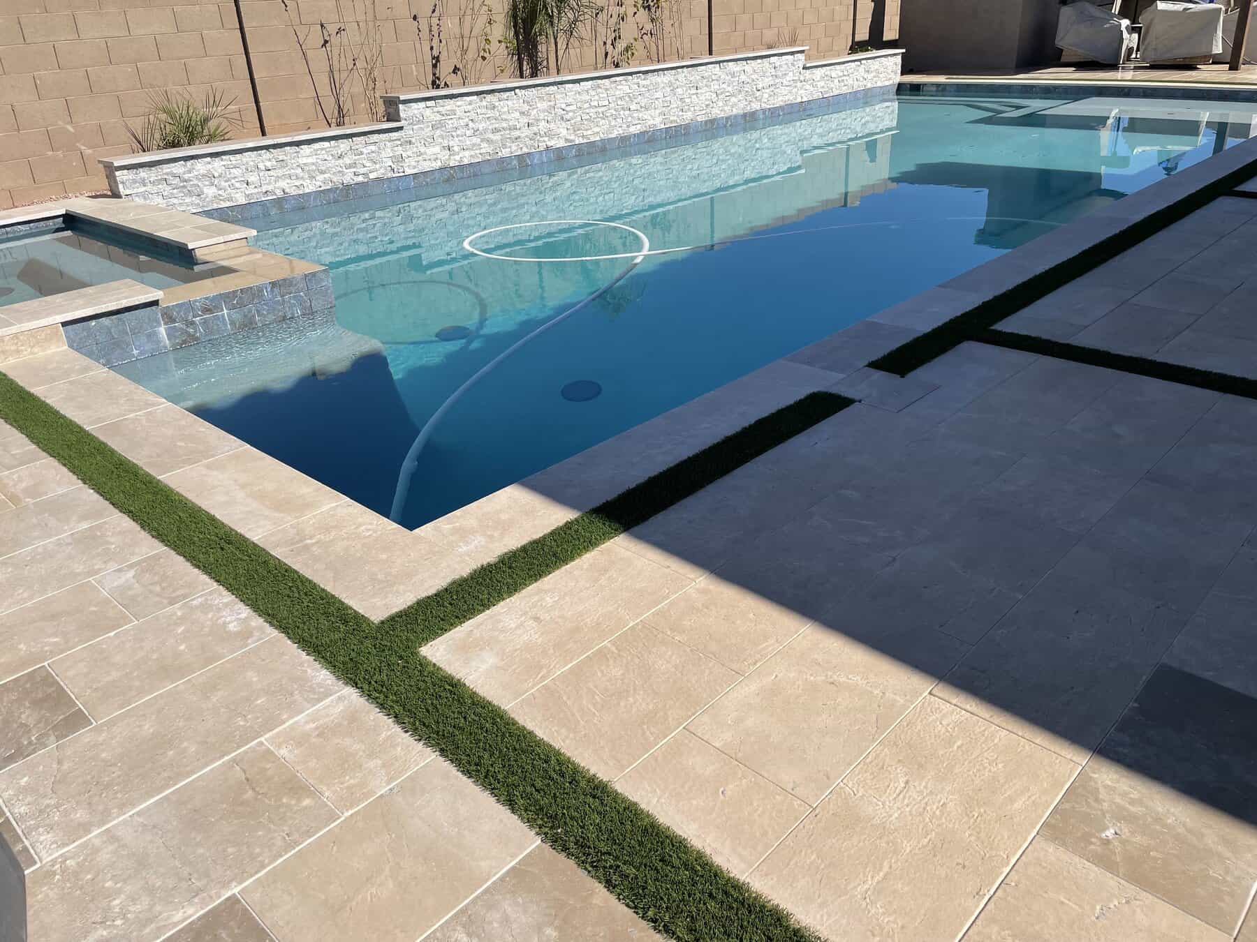 A Texas backyard with artificial grass, bordered by rocks and gravel and enclosed by a tan brick wall. Trees and bushes line the fence, and a storage bench sits against the wall under a clear blue sky.