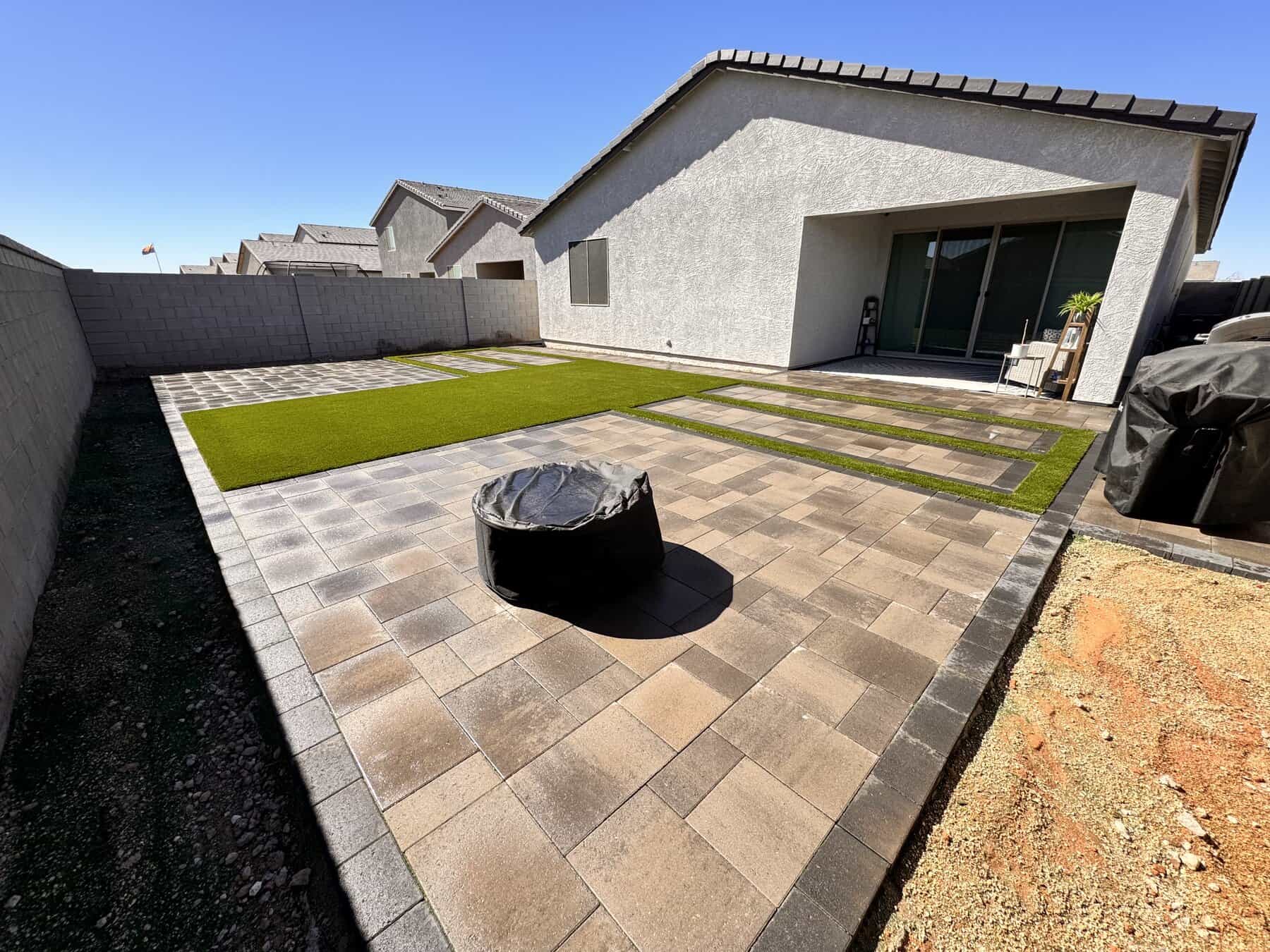 A backyard putting green with artificial turf, golf flags, large rocks, a white sand trap, and lush landscaping, all set against a tall tan wall and adjacent to a nearby patio area.