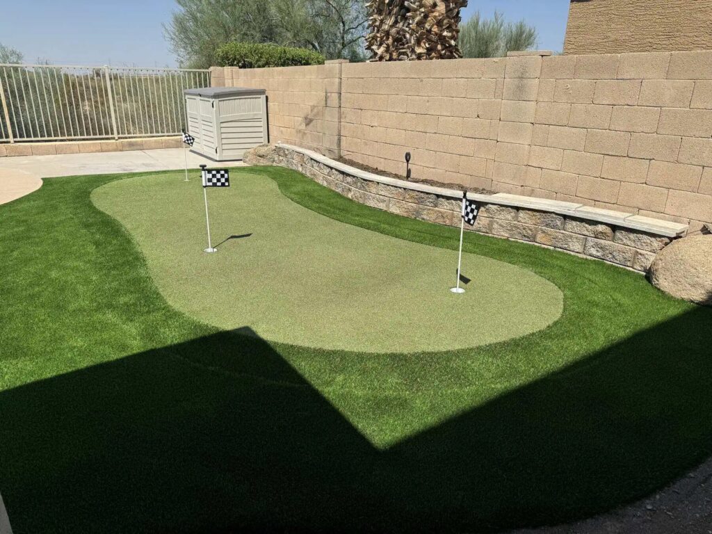 A backyard putting green with two golf holes, each marked by a black-and-white checkered flag, is surrounded by lush artificial turf, stone edging, and a tan brick wall. A storage shed sits in the corner.