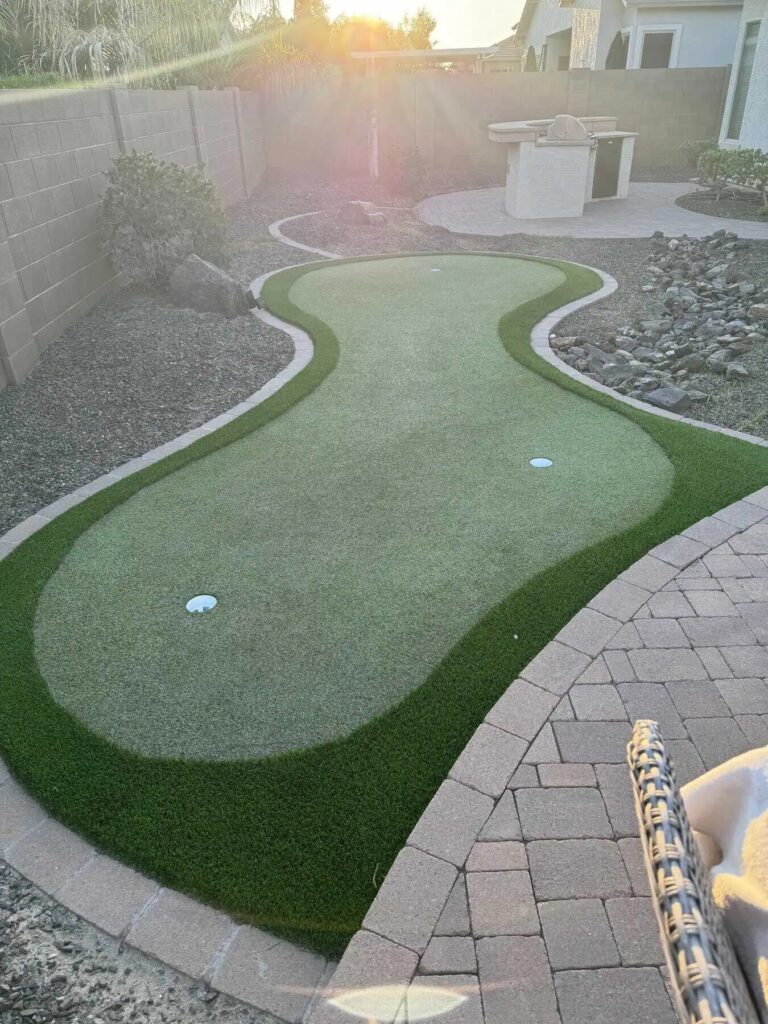 A backyard featuring a kidney-shaped putting green bordered by bricks, surrounded by rocks, lush plants, and a barbecue area, with sunlight streaming in from above.