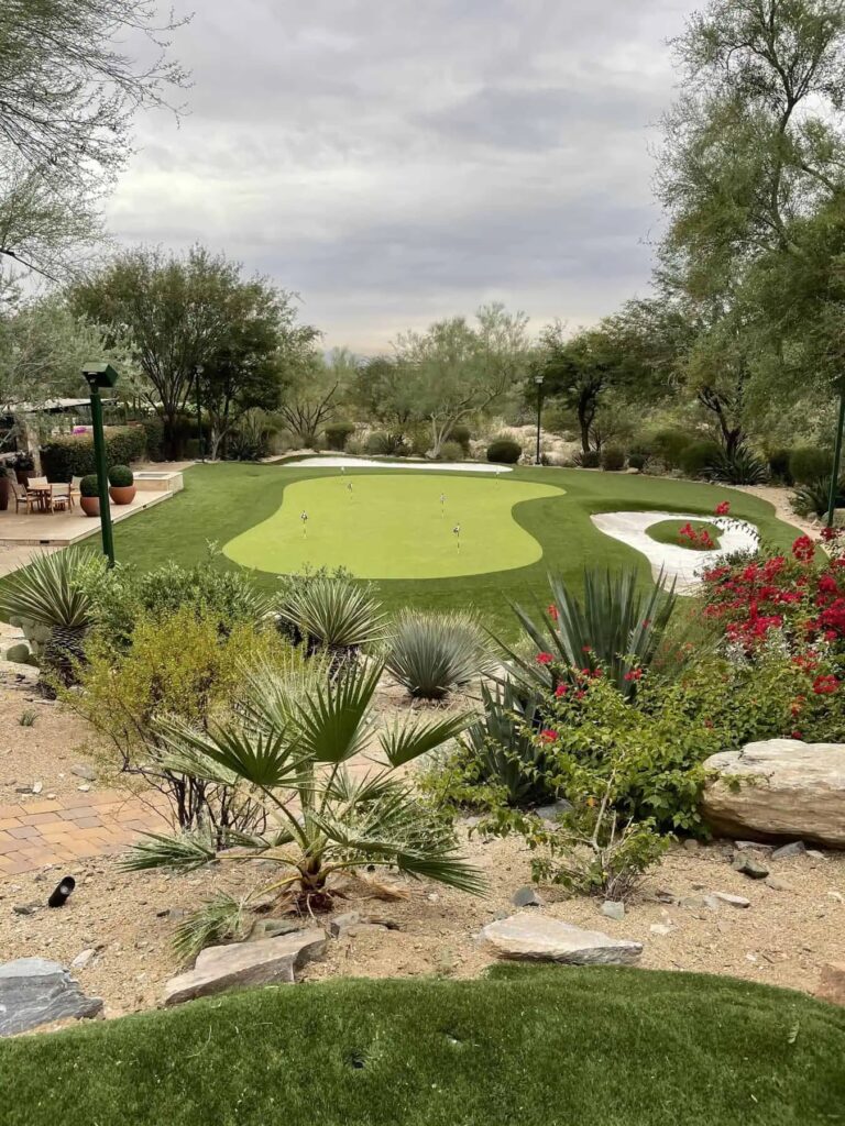 A landscaped backyard features a small putting green surrounded by desert plants, bushes with red flowers, rocks, a sand trap, and patio seating under cloudy skies.