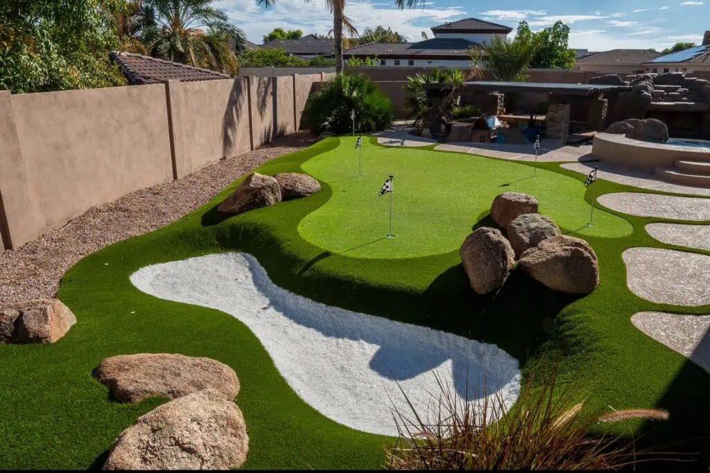 A backyard putting green with artificial turf, golf flags, large rocks, a white sand trap, and lush landscaping, all set against a tall tan wall and adjacent to a nearby patio area.