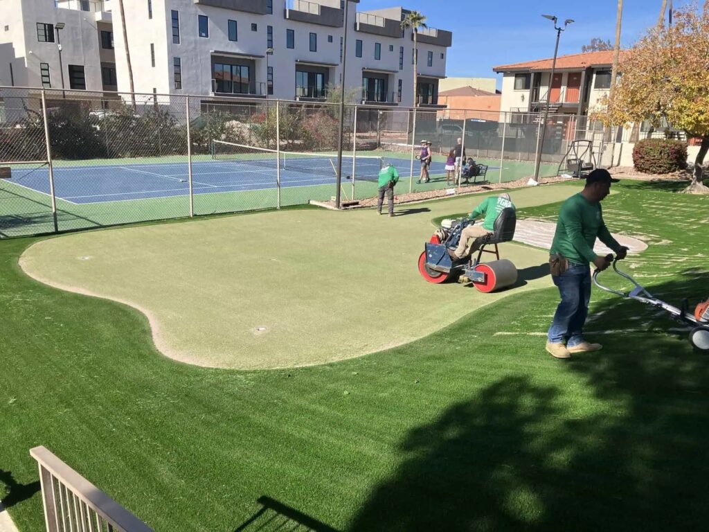 Workers expertly install artificial turf on a putting green near a tennis court, with apartment buildings in the background and clear blue skies overhead.