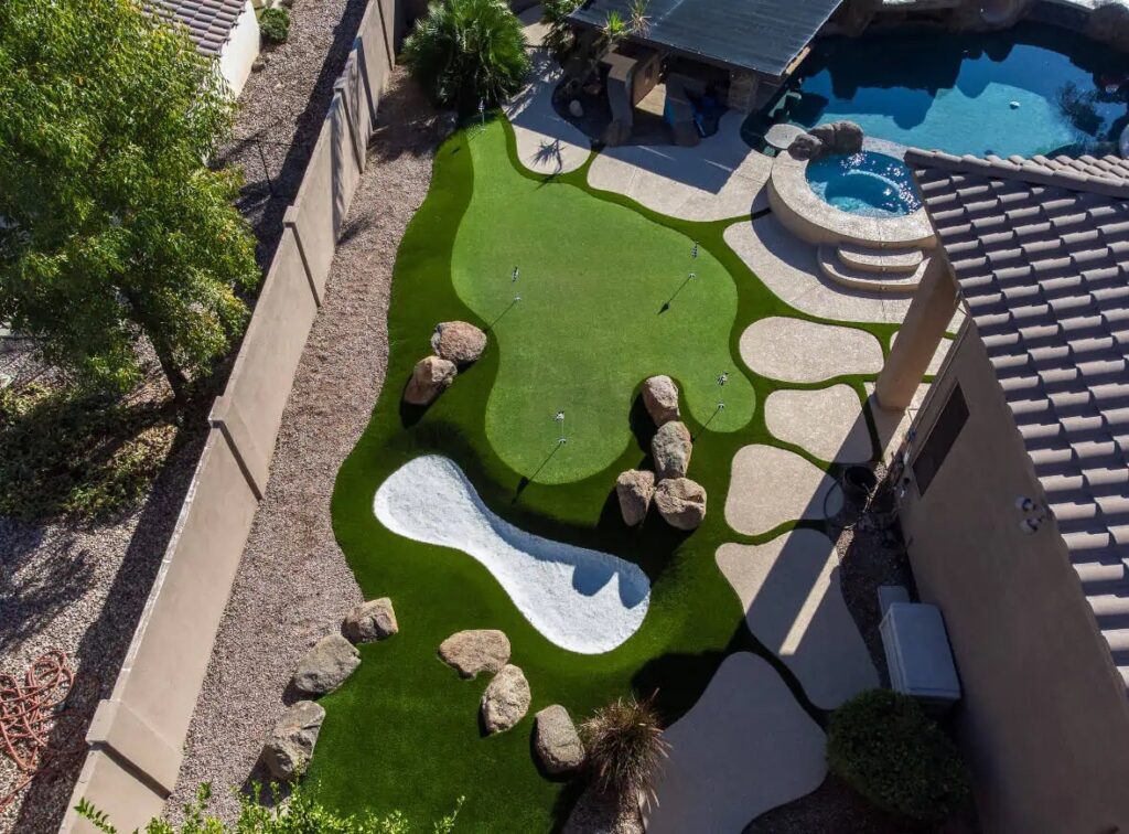 Aerial view of a backyard featuring a putting green, artificial turf, sand trap, rocks, desert landscaping, and a swimming pool with a shaded patio area.
