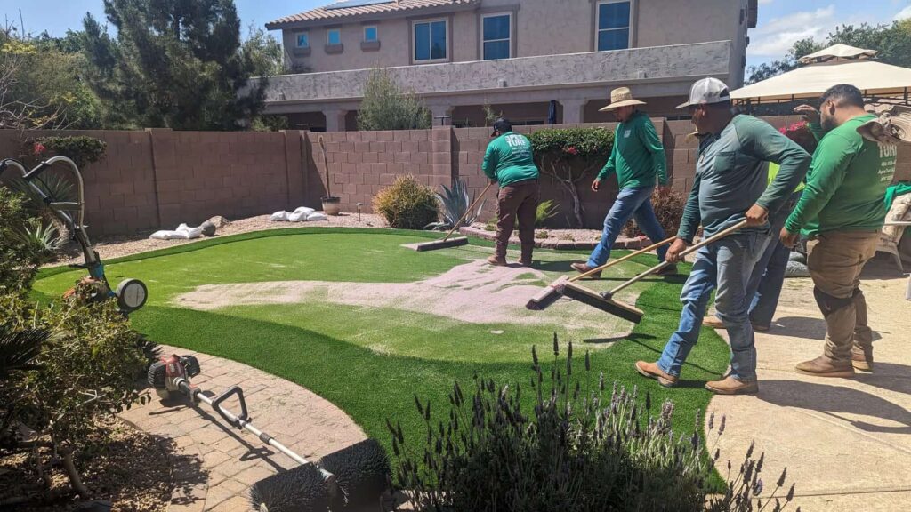 Four workers in green shirts spread sand over an artificial putting green in a backyard. A brick house and garden plants are visible, with tools and equipment scattered on the ground nearby.