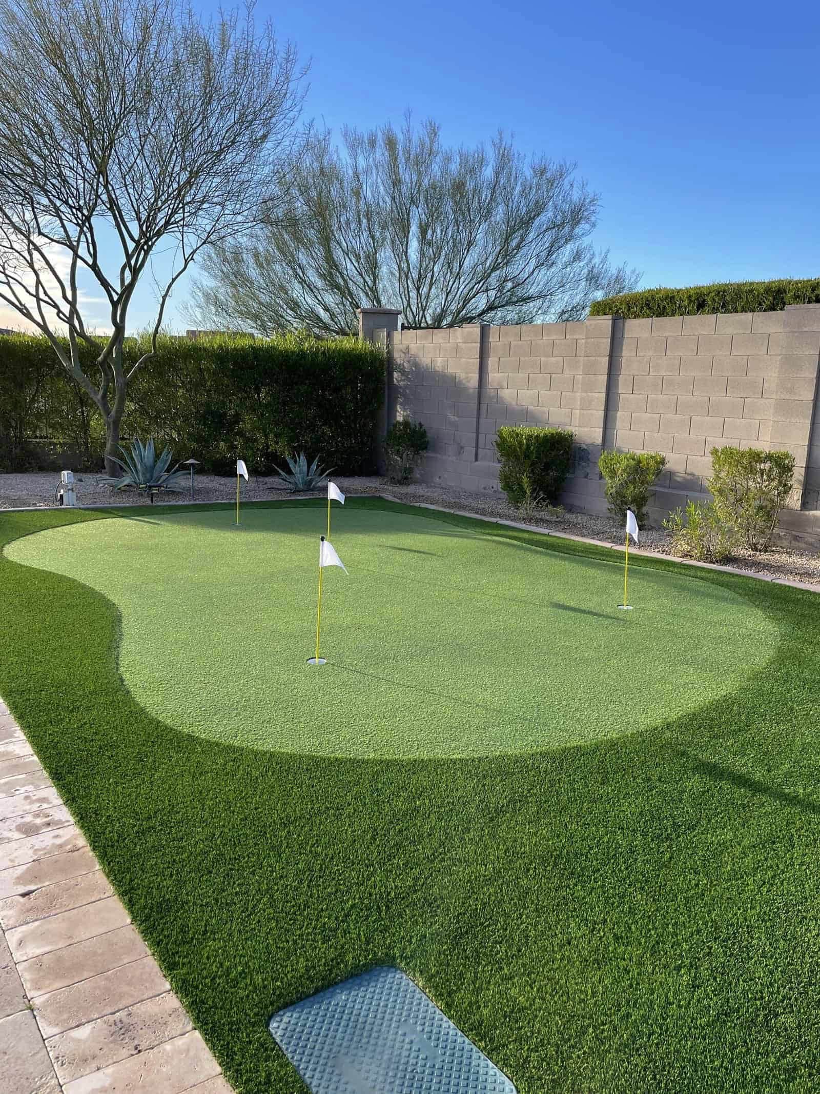 A backyard putting green with four small flag poles, surrounded by artificial grass, desert plants, and a beige brick wall creates the perfect spot to practice your golf short game under a clear blue sky.