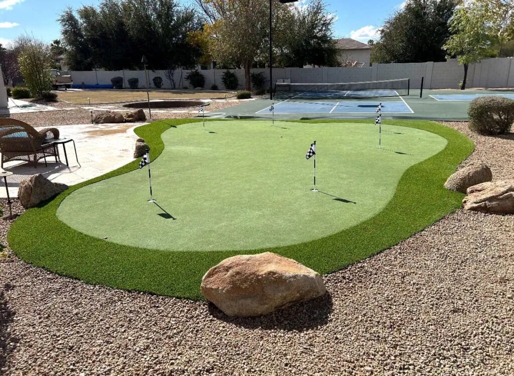 A backyard putting green features four golf holes with flags, surrounded by artificial turf, rocks, and gravel. In the background, a tennis court and trees complete this inviting outdoor space.