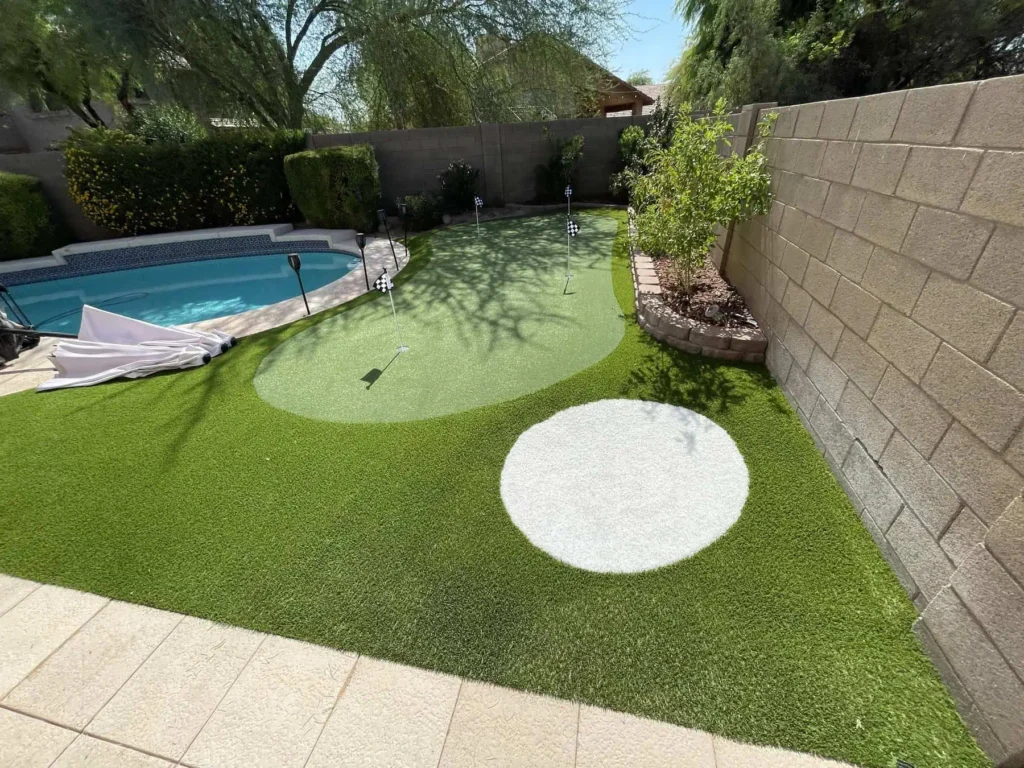 A backyard features a swimming pool and a custom putting green with artificial turf. Golf flags, a round white area mimicking a golf ball, and landscaped plants and trees line the wall for a lush, resort-style vibe.