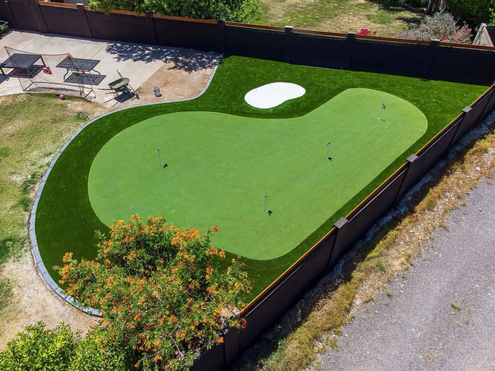 A Texas backyard with artificial grass, bordered by rocks and gravel and enclosed by a tan brick wall. Trees and bushes line the fence, and a storage bench sits against the wall under a clear blue sky.